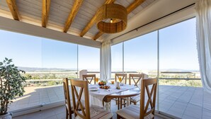 Un coin repas avec une table dressée pour quatre personnes, entourée de chaises en bois, de grandes fenêtres offrant une vue sur le paysage et un luminaire suspendu.