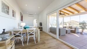Une pièce spacieuse avec une table à manger en bois, des chaises blanches, une grande fenêtre donnant sur l’extérieur et un ventilateur de plafond.