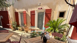 An outdoor patio with wooden tables and chairs, a small garden with potted plants, and a building with red shutters and a sign reading 'La Bastide.' - Charming Provençal studio with peaceful patio. Alpilles (Eyragues)