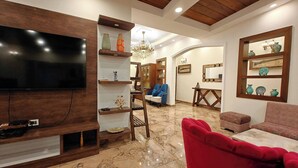 A living room with a TV, wooden shelves, a red sofa, a chandelier, and a decorative mirror. - The Paradise - Luxury 3BR Villa Near the Airport in Dehradun (Rishikesh)
