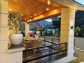 A covered outdoor seating area with wicker furniture, potted plants, and a view of a mountain and bridge in the background. - The Paradise - Luxury 3BR Villa Near the Airport in Dehradun (Rishikesh)
