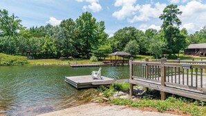 A lakeside dock with a wooden pier, a covered seating area, and a building in the background.