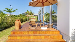 A wooden deck with a chair, table, and umbrella, overlooking a garden and a modern building.