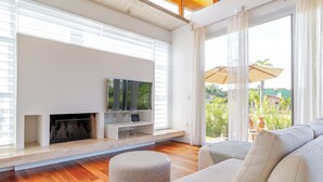 A modern living room with a fireplace, a flat-screen TV, and a large window with a view of greenery.