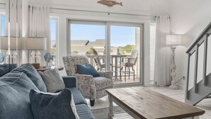 A spacious living area with a large sofa, patterned armchair, and a wooden coffee table. A fish decoration hangs from the ceiling, and there is a view of a patio with outdoor furniture.