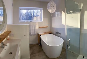 Un baño moderno con una bañera independiente, un box de ducha de vidrio, un tocador de madera y un ventanal que proporciona luz natural.