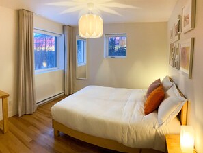Un dormitorio con cama, mesita de noche, lámpara colgante y dos ventanas.