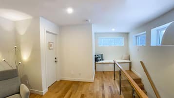 A modern, well-lit hallway with a wooden floor, a grey armchair, a bed with a geometric-patterned pillow, and a window providing natural light.