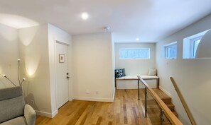 Un pasillo moderno y bien iluminado con suelo de madera, un sillón gris, una cama con almohada de estampado geométrico y un ventanal que proporciona luz natural.