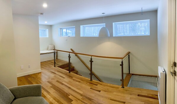 Un interior moderno con escalera de madera, un pequeño rincón de descanso y ventanas grandes.