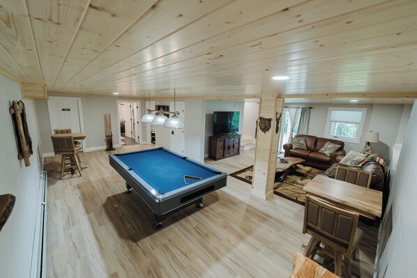 Un vaste espace de vie comprenant une table de billard, un ensemble de canapés, un téléviseur et un plafond en bois.