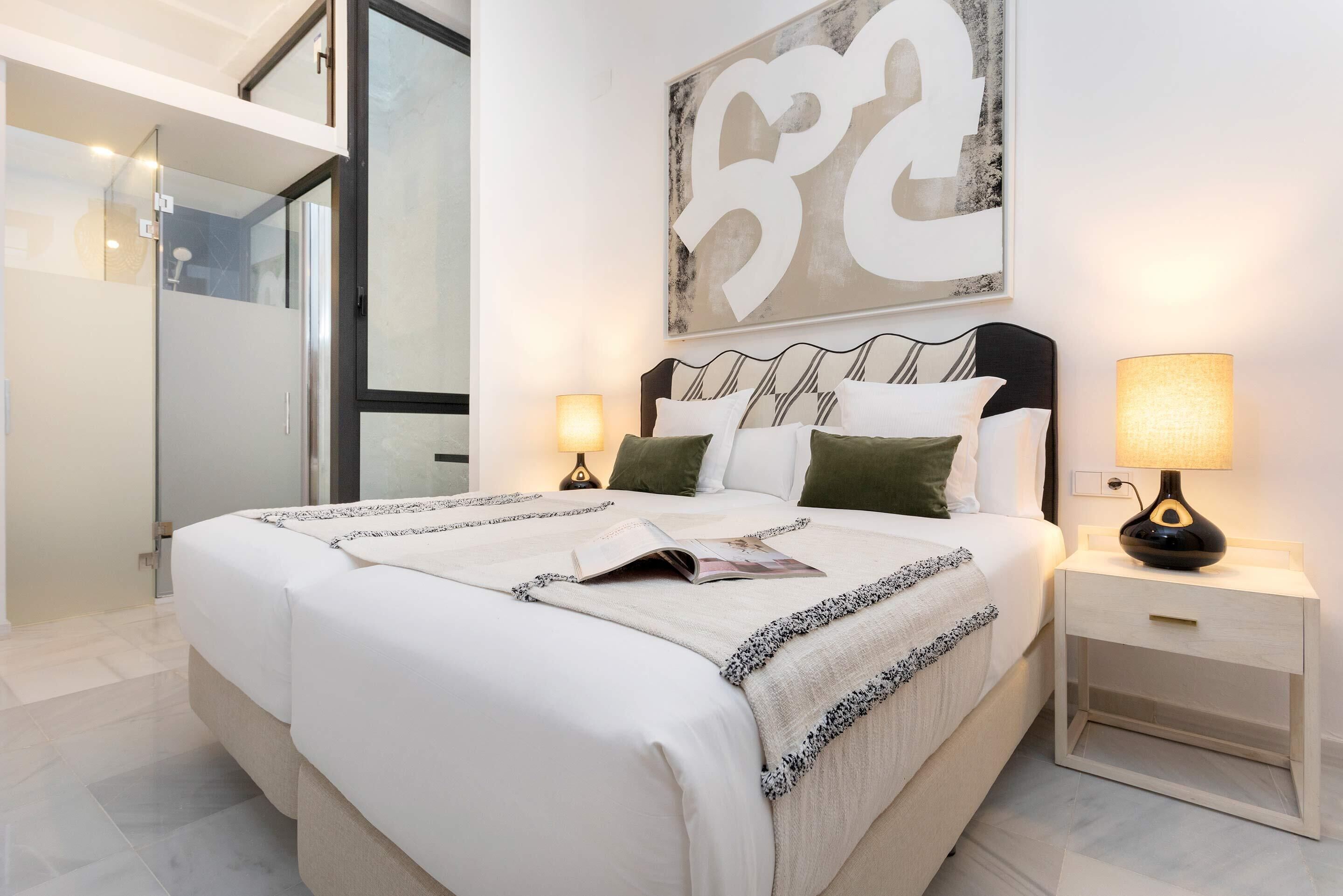 Une chambre moderne avec un grand lit, deux tables de chevet avec des lampes, une cabine de douche en verre et un grand tableau abstrait accroché au mur.