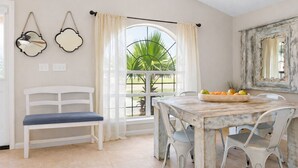 Un coin repas avec une table en bois rustique, des chaises blanches et un banc. Une fenêtre avec des rideaux laisse entrer la lumière naturelle et offre une vue sur la verdure à l’extérieur.