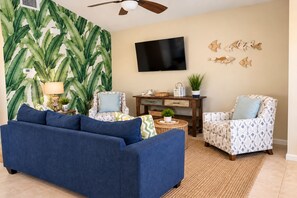 Una sala de estar con un sofá azul, sillones estampados, un televisor de pantalla plana, arte mural decorativo de peces y una planta grande.
