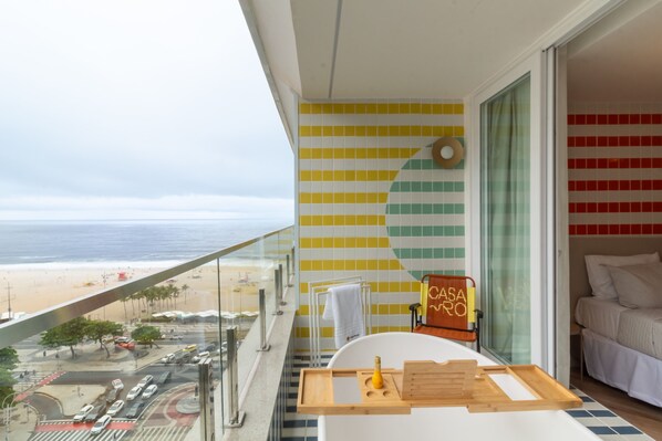 A balcony with a view of the beach and ocean, a bathtub, and a wooden tray with a bottle.