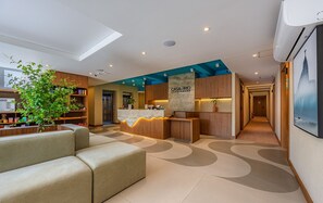 A modern hotel lobby with a reception desk, a seating area, and a kitchen visible in the background.