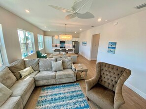A spacious living room with a sectional sofa, a tufted armchair, and a dining area in the background. - Nautical Nest - Quick Walk to Beach (Fort Myers Beach)