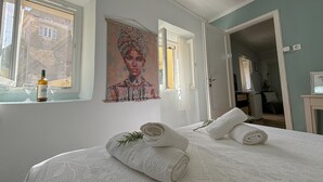 A bedroom with a bed, two rolled towels, a painting, and a bottle of wine on the windowsill.