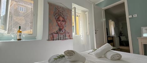 A bedroom with a bed, two rolled towels, a painting, and a bottle of wine on the windowsill.