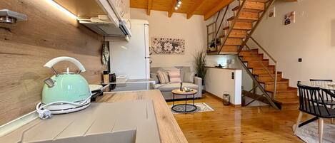 Una cocina moderna con un lavamanos, hervidor y suelo de madera. Se ve una escalera que sube a una zona estilo altillo con un sofá y una mesita.