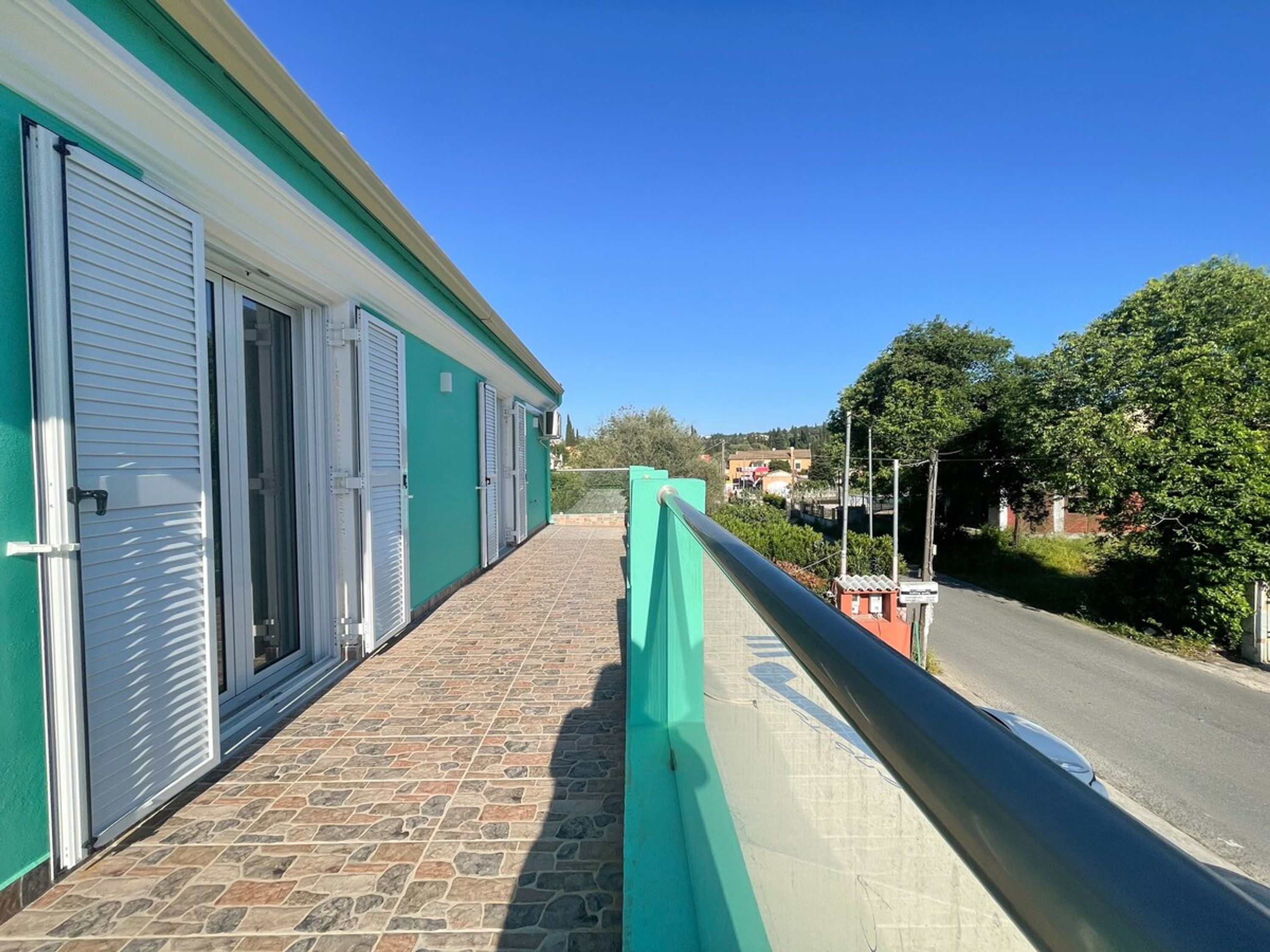 Un bâtiment à l’extérieur verdoyant, doté d’un balcon pavé de pierres et offrant une vue sur une route et la verdure environnante.