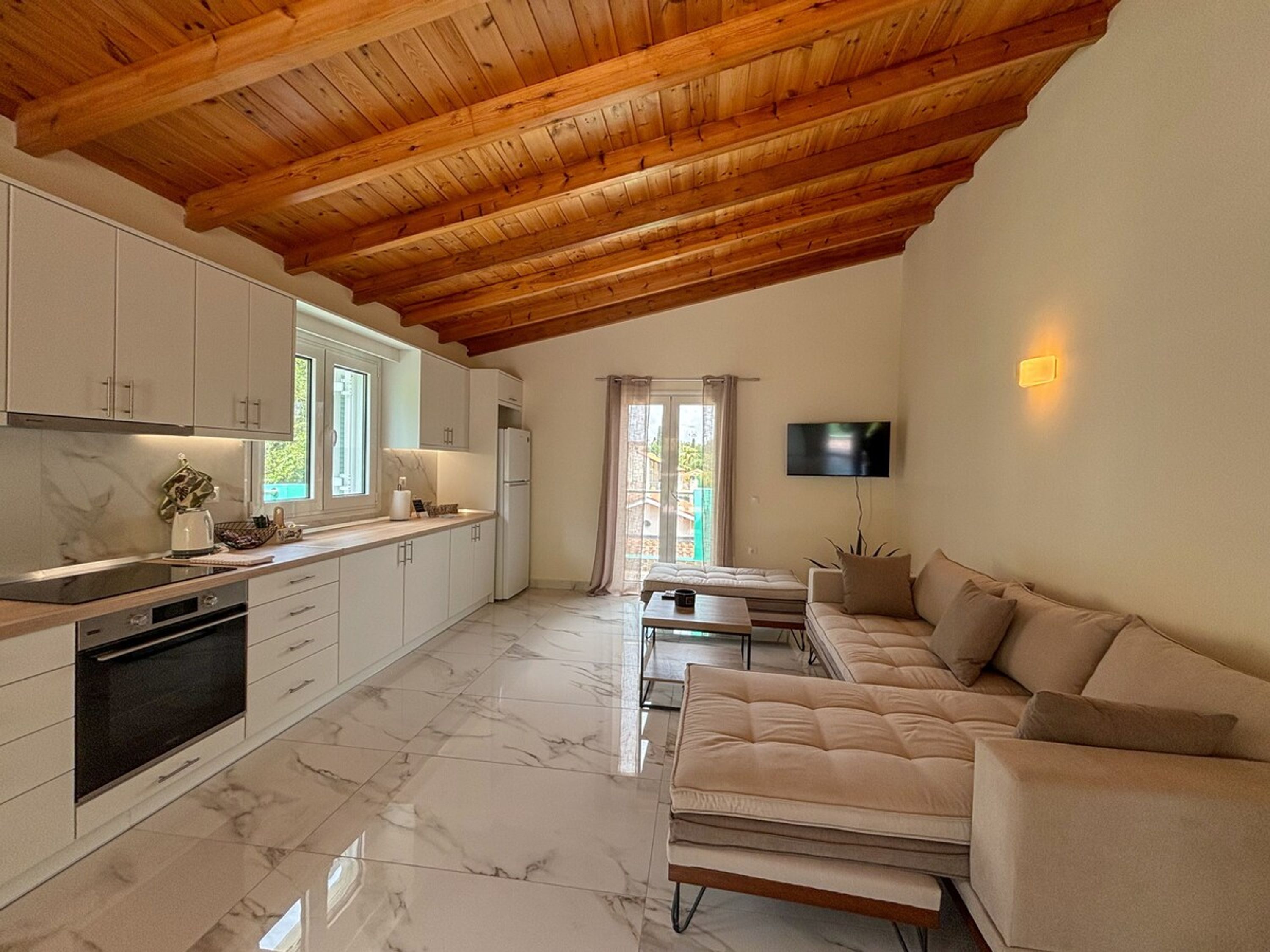 A modern kitchen with marble flooring, wooden ceiling beams, and a dining area with a sofa and table.