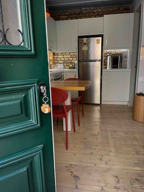 A compact kitchen with a refrigerator, a stove, and a sink. There are two red chairs and a wooden table. The floor is wooden, and the walls have a brick pattern. - Kukuvayia s Nest-centre Old Town Corfu (Corfu)