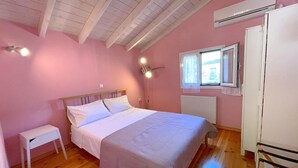 A bedroom with a wooden floor, a bed with white bedding, a small white bedside table, a window with a view, and an air conditioning unit on the wall. - Ourano -gastouri Village Center (Corfu)
