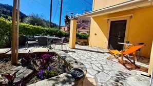 A patio area with stone flooring, outdoor furniture, and a garden bed with purple flowers. - Ourano -gastouri Village Center (Corfu)