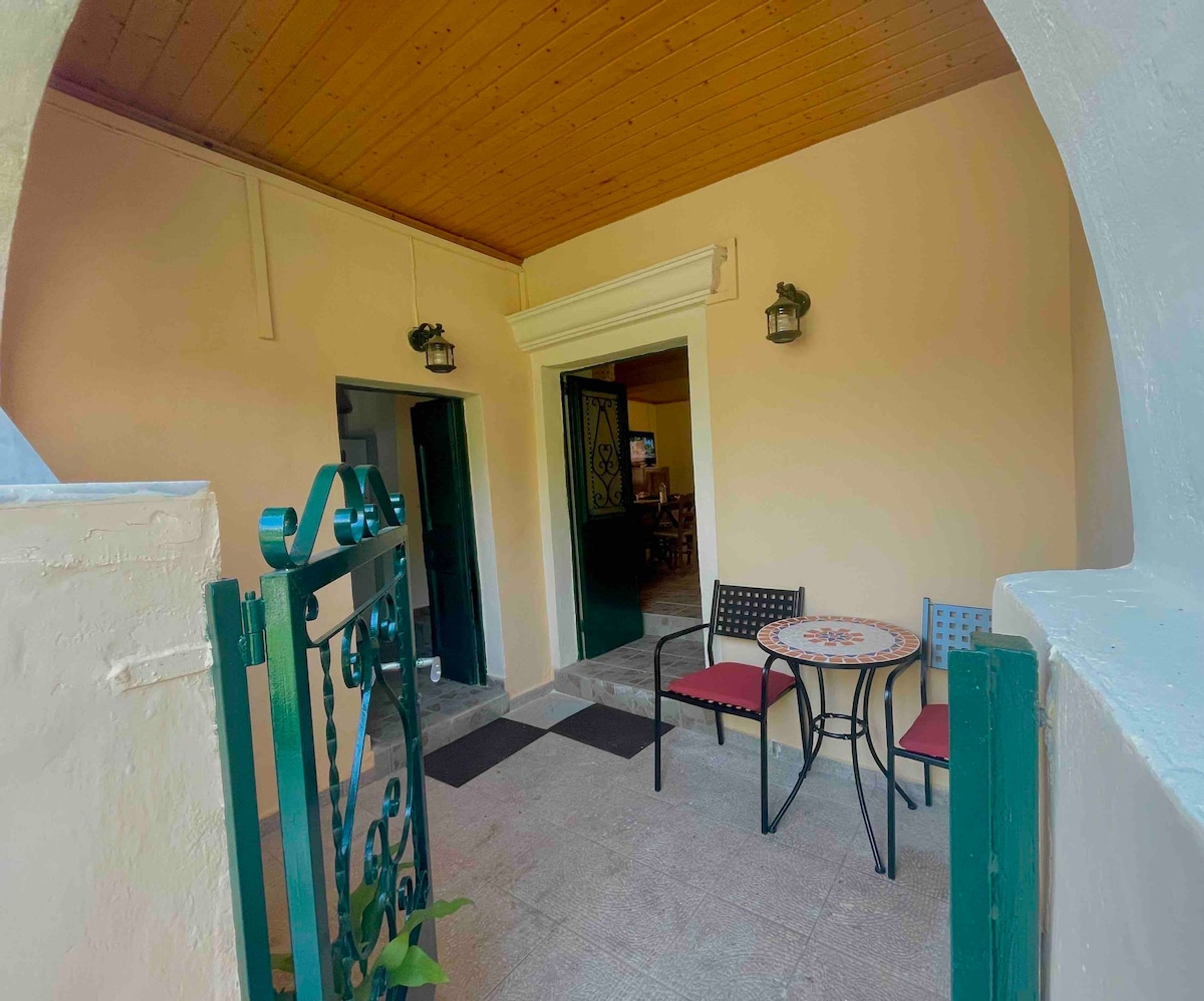 A covered outdoor seating area with a table and chairs, a metal gate, and a door leading to an interior space.
