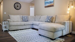 A living room with a light-colored sectional sofa, patterned throw pillows, a decorative rug, a wall clock, a floor lamp, and a painting. - Charming 3-bedroom home in Fayetteville: 5 min from Base (Fayetteville)