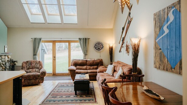 A spacious living room with a large window, a brown leather sofa, a wooden dining table, and a rustic decor theme. - Ski-In/Ski-Out Townhouse at Tenney Mountain Resort (Plymouth)