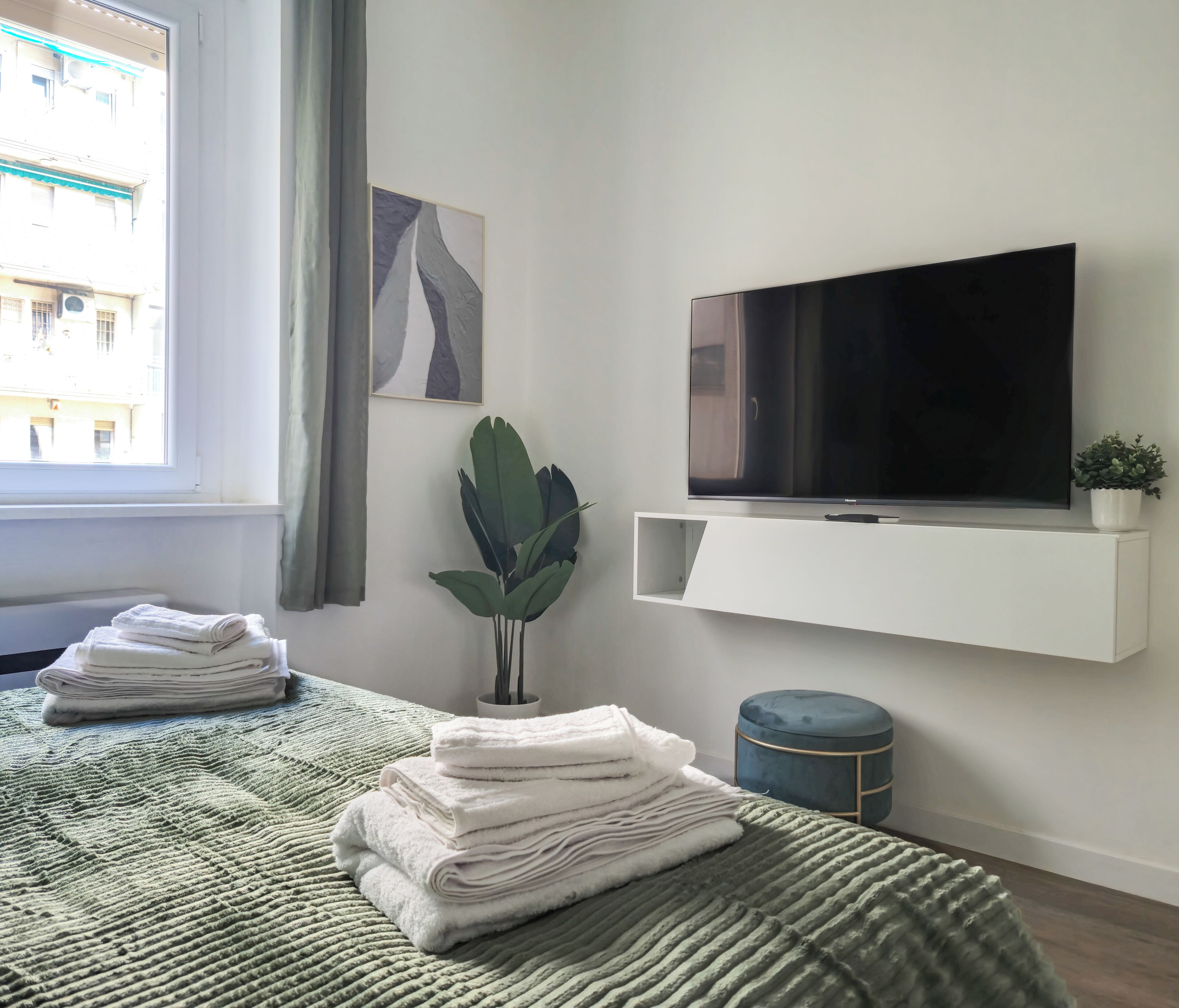 A modern hotel room with a bed, a TV mounted on a wall, a small potted plant, and a window with curtains.