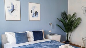 A bedroom with a large bed, two wall-mounted artworks, a potted plant, and a nightstand with a lamp. - Morgagni Suites (Bologna)