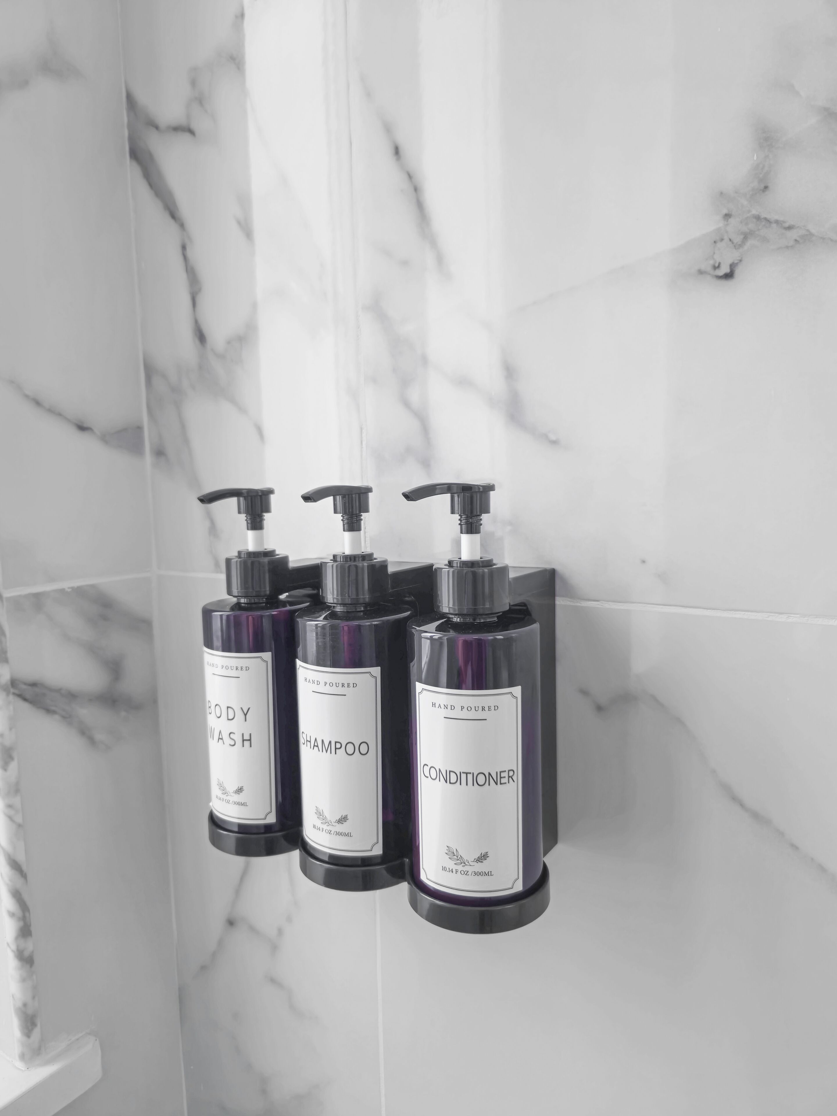 Tres botellas de gel de baño, champú y acondicionador hechas a mano, colocadas en una pared con diseño de mármol.