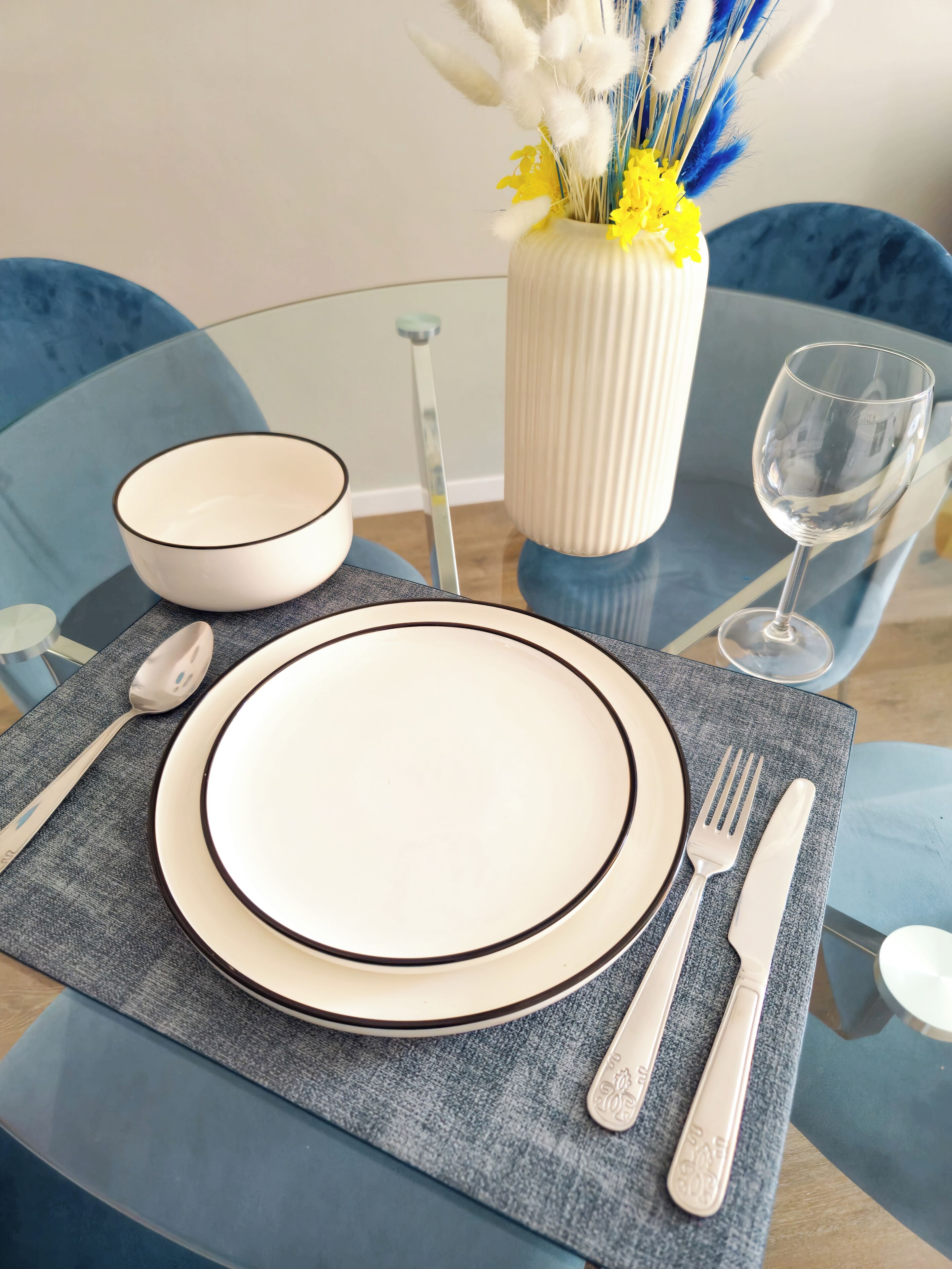 A dining table set with a white plate, bowl, cutlery, and a vase with yellow flowers.