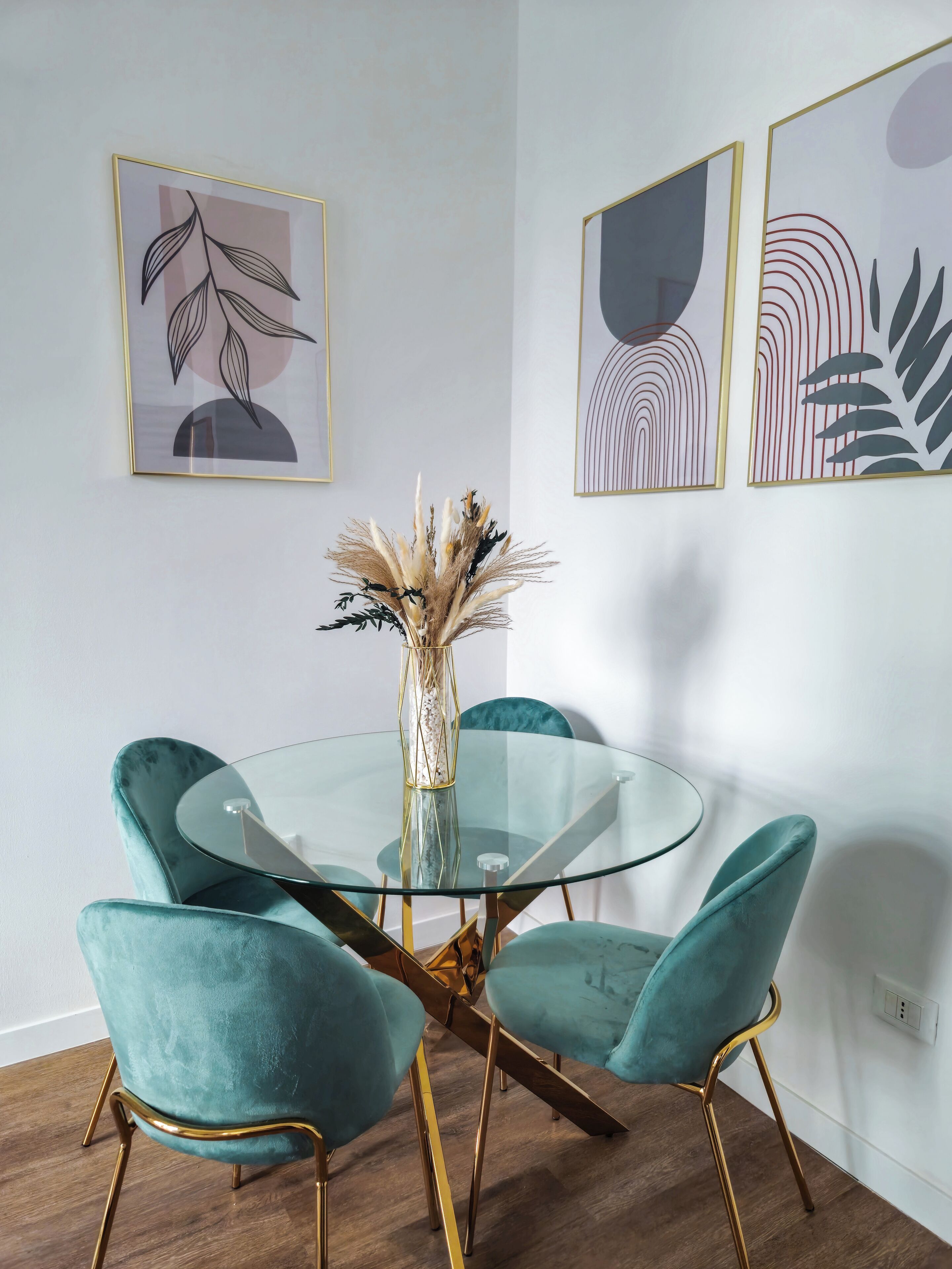 A dining area with a glass table surrounded by teal chairs with gold legs, a vase with dried plants, and abstract art on the walls.