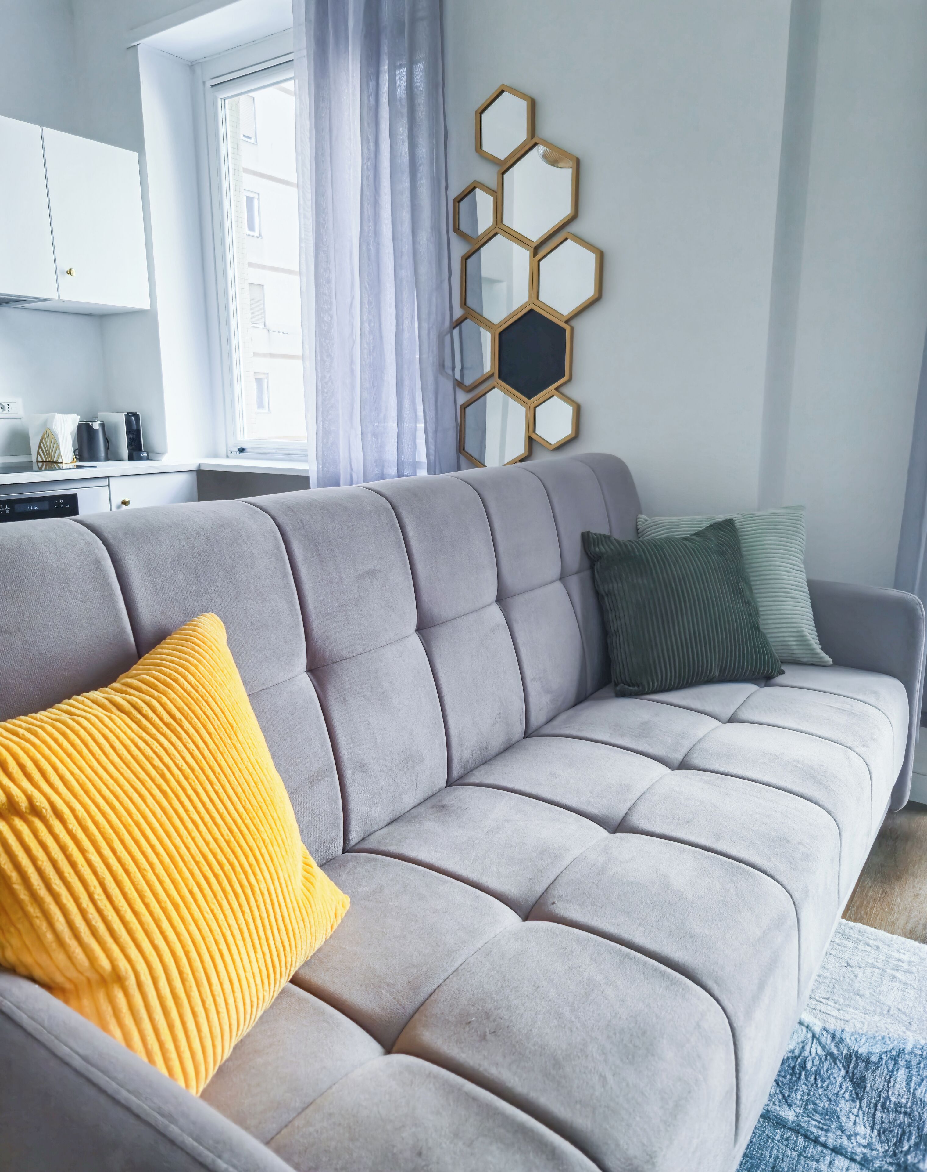 A modern living room with a grey sofa, a yellow pillow, and a green pillow. There is a hexagonal mirror on the wall and a window with curtains.