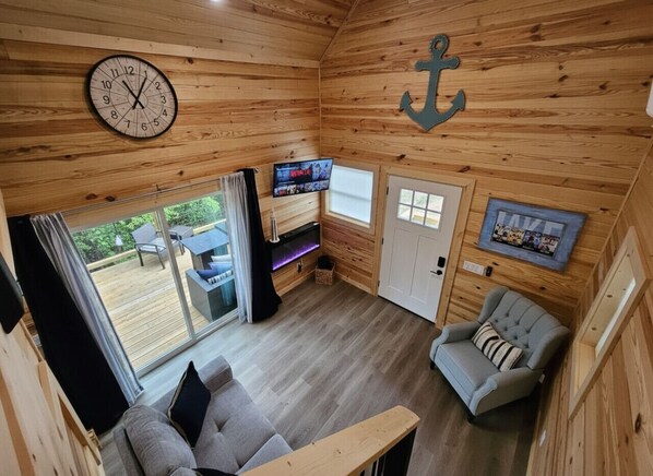 Acogedor interior de cabaña con detalles de madera, un reloj, un televisor, una chimenea y vistas al exterior.