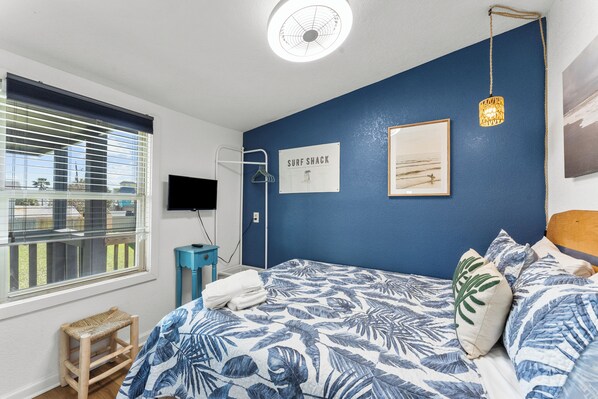 Un dormitorio con cama, ventana con persianas, televisor montado en la pared, cartel que dice SURF SHACK, y una lámpara colgante.