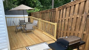 Een houten terras met een barbecue, buitentafel, stoelen en een overdekte parasol.
