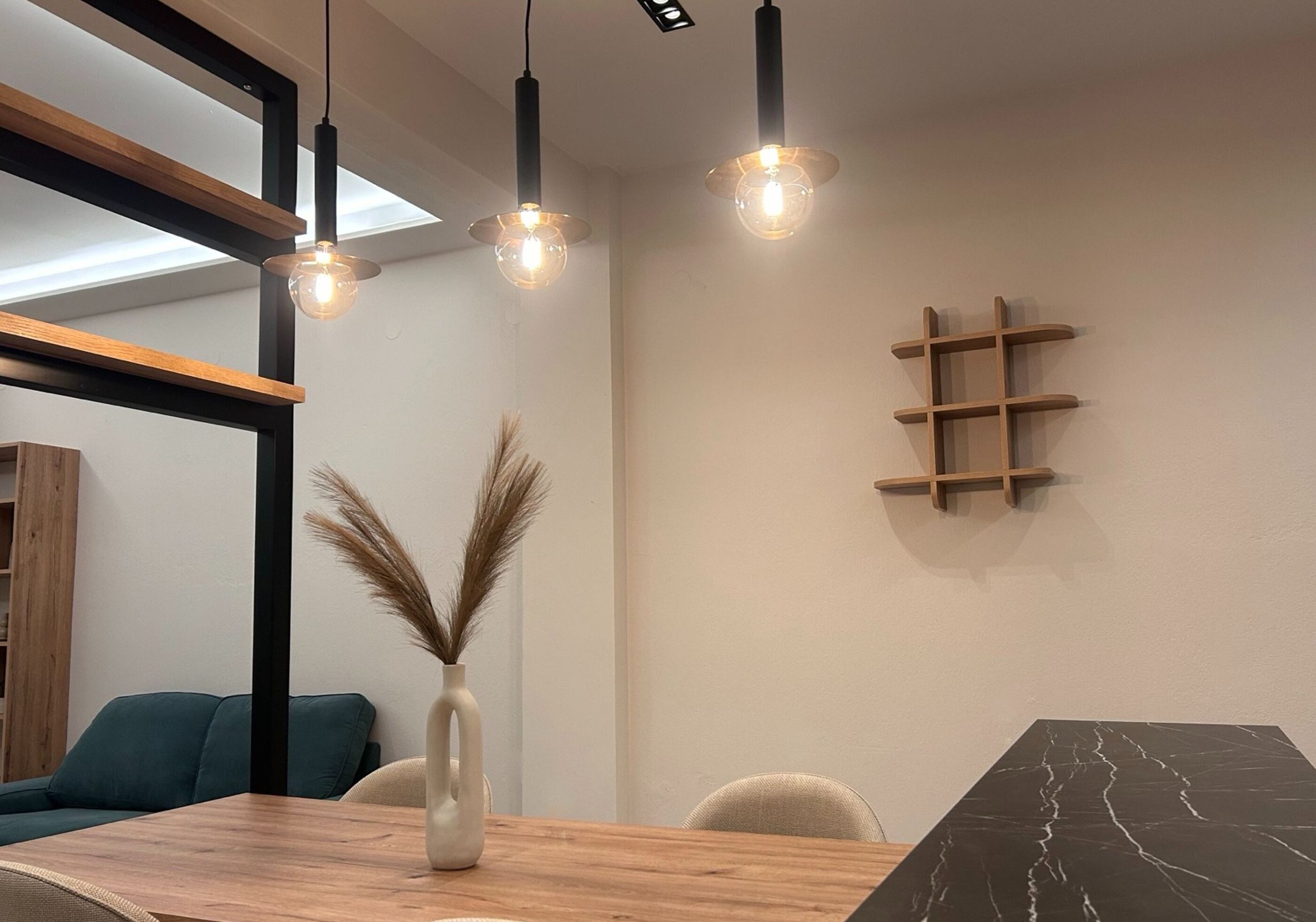 Uma área de refeições com mesa de madeira, cadeiras, um vaso com plumas de capim-do-campo, luminárias pendentes e um detalhe em mármore preto.