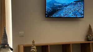 A living room with a wooden table, a small Christmas tree, a gnome, and a television displaying a snowy landscape. - Abete Luxury Apartments (Metsovo)