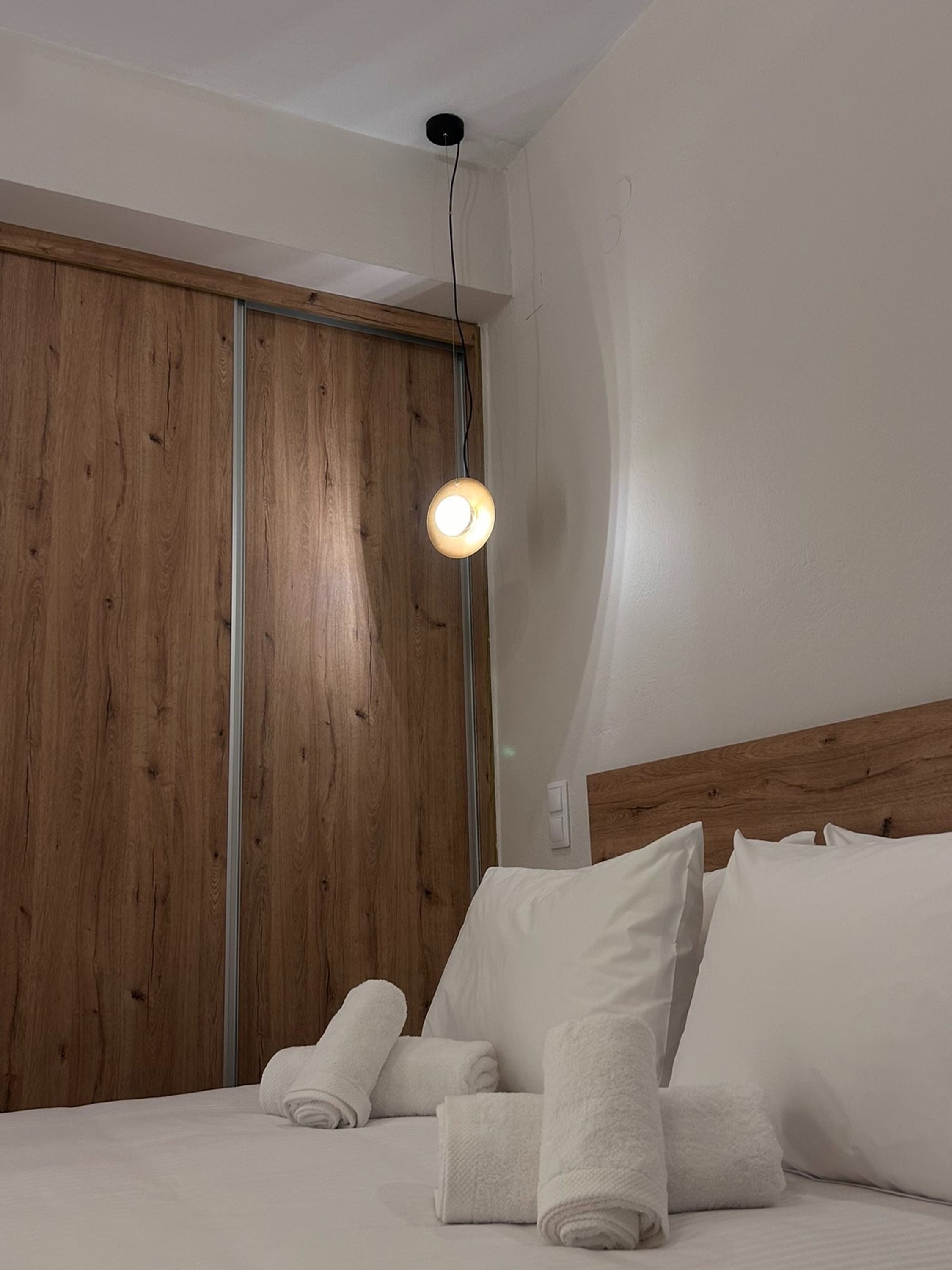 Quarto com guarda-roupa de madeira, cama com roupa de cama branca e luminária pendente.