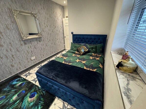 A bedroom with a tufted headboard, a peacock-themed bedspread, a decorative mirror, and a window with blinds. - Central Location - Apartment - Sleeps 2 - Parking (Gloucester)