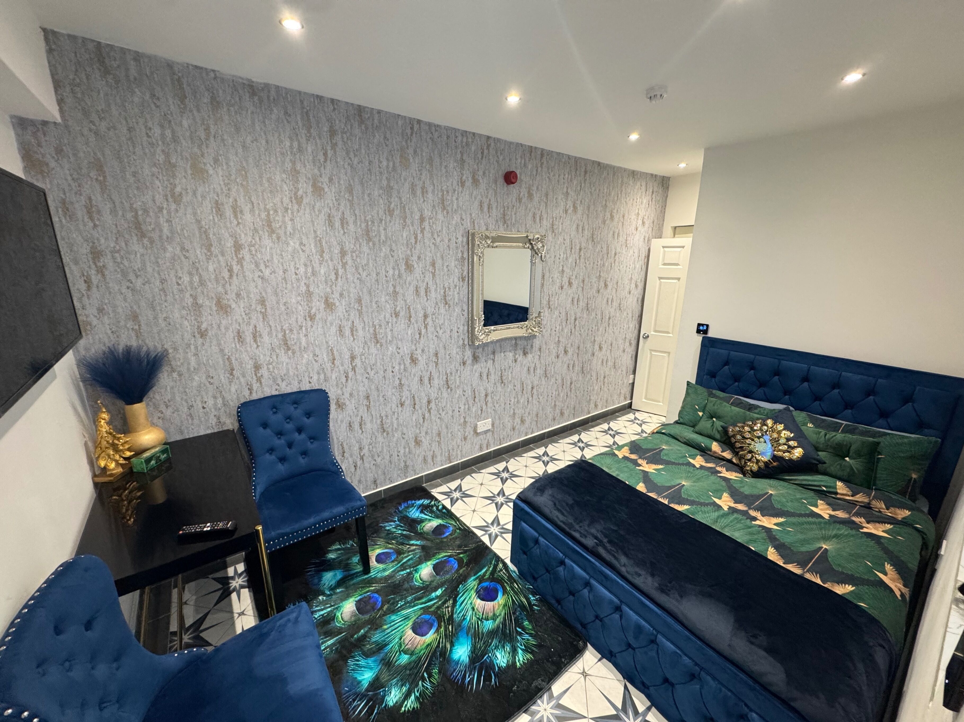 A bedroom with a peacock-themed rug, a tufted blue chair, a bed with a blue headboard, and a mirror on the wall.