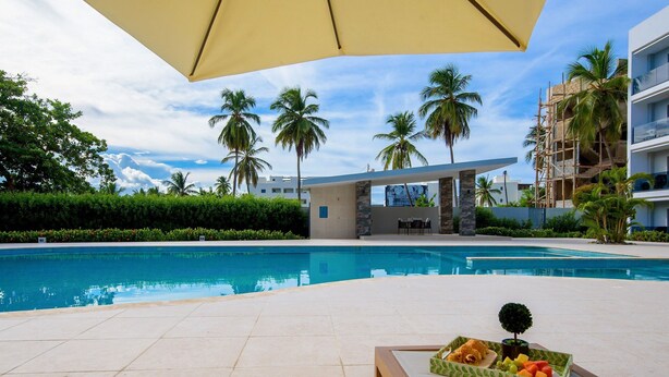 A poolside area with a lounge chair, a table with a fruit tray, and a view of the pool and palm trees. - Bran new condo with pool access (Bavaro)