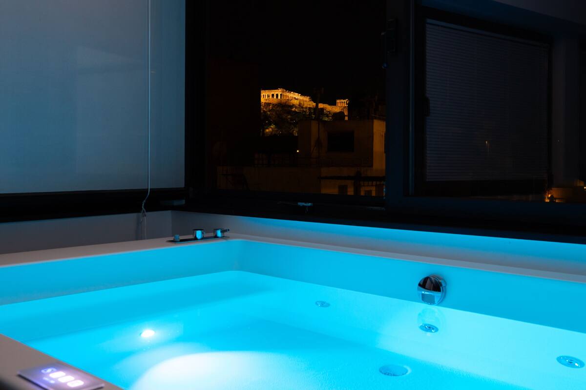 A modern bathroom with a large bathtub, a visible control panel, and a window with a view of a building at night.