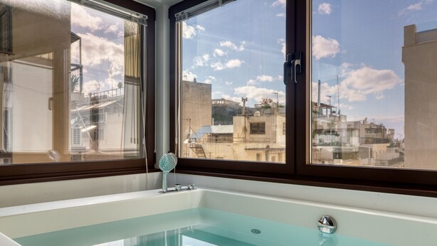 A modern bathroom with a glass-enclosed bathtub, large windows offering a view of the cityscape, and a visible showerhead. - GSM Elevated Luxury Suites (Athens)