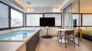 A modern hotel room with a large window, a flat-screen TV, a small table, and a bed. - GSM Elevated Luxury Suites (Athens)
